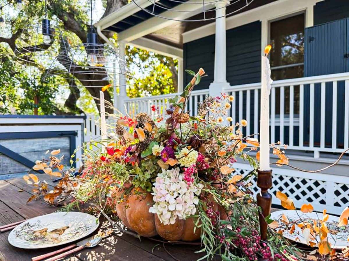 A rustic outdoor table features a Dried Flower Pumpkin Centerpiece, overflowing with vibrant blooms and foliage. Plates and utensils are set nearby, with a candle and a white house with blue shutters in the background.
