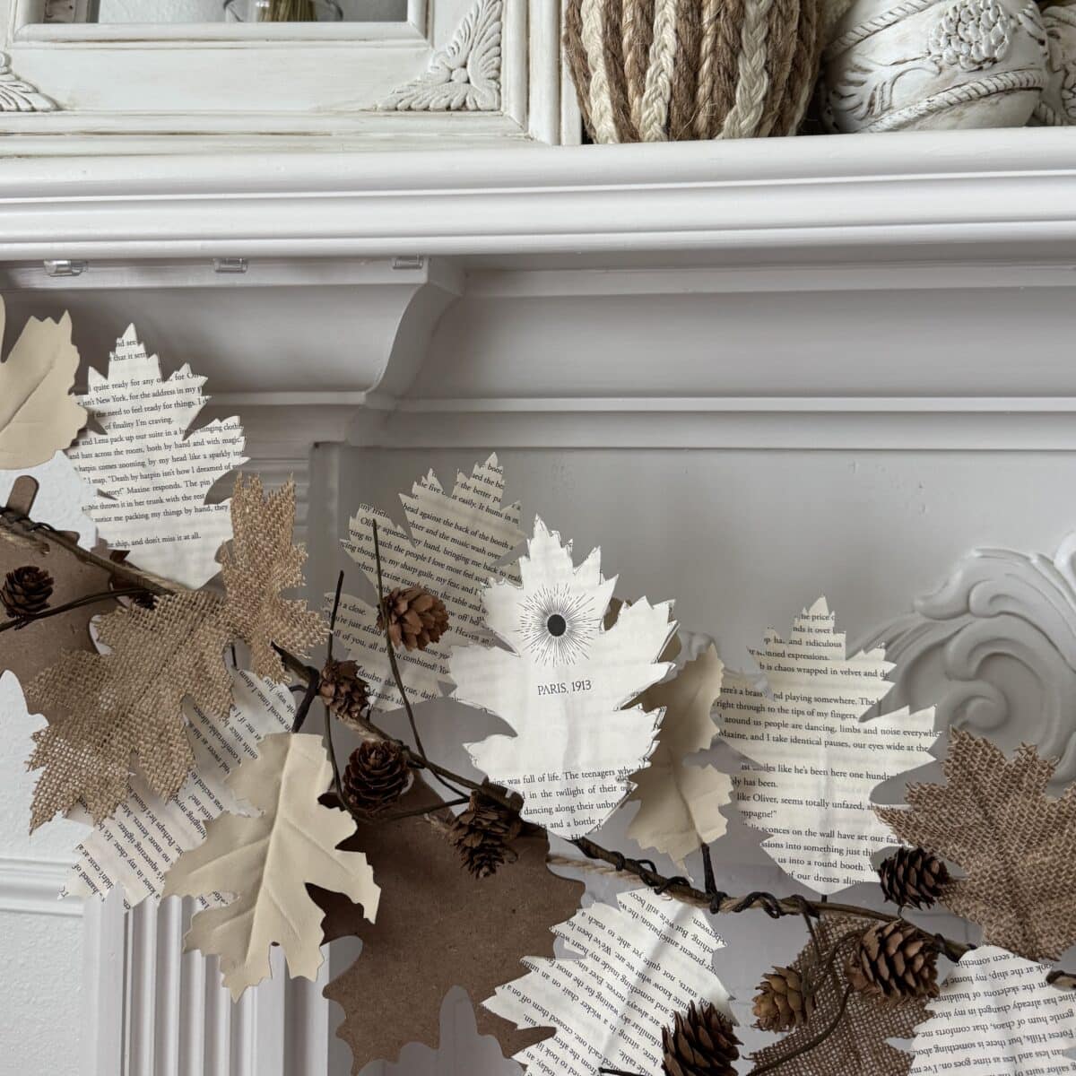 A garland of artificial leaves made from book pages and burlap drapes across a white ornate mantel, with pinecones attached. Decorative woven and ceramic objects sit on the shelf above.