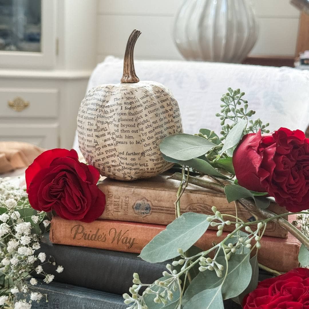 A small pumpkin covered in book pages sits atop stacked vintage books, including one titled "Pride's Way," with red roses and greenery arranged around it on a cozy, light-colored chair.