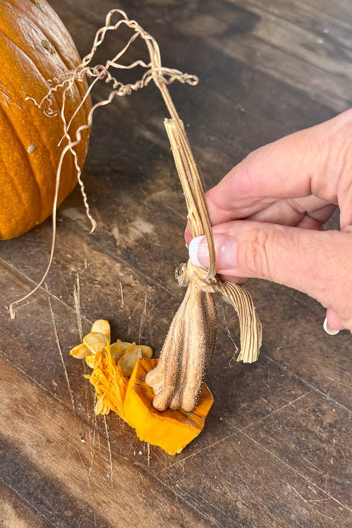 A hand holds a small broom made from pumpkin stem, seeds, and raffia, resting on a wooden surface next to a partially visible pumpkin.