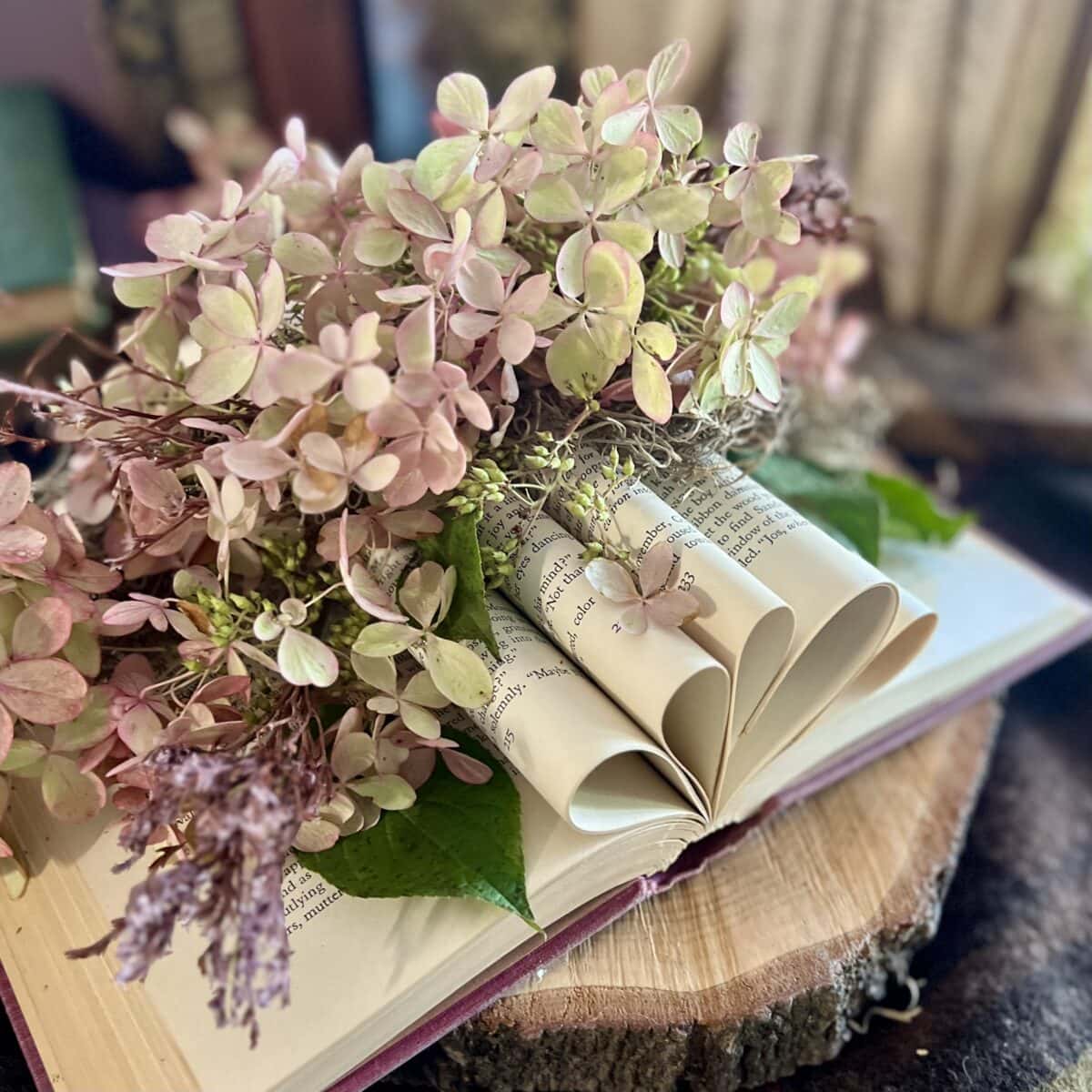 An open book with its pages artistically folded into heart shapes, adorned with pink and green hydrangea flowers and greenery, resting on a wooden slice.