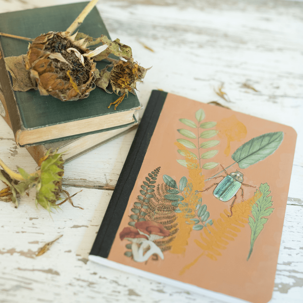 A botanical-themed notebook with fern and beetle illustrations lies on a white wooden surface next to two vintage books and a few dried flowers.