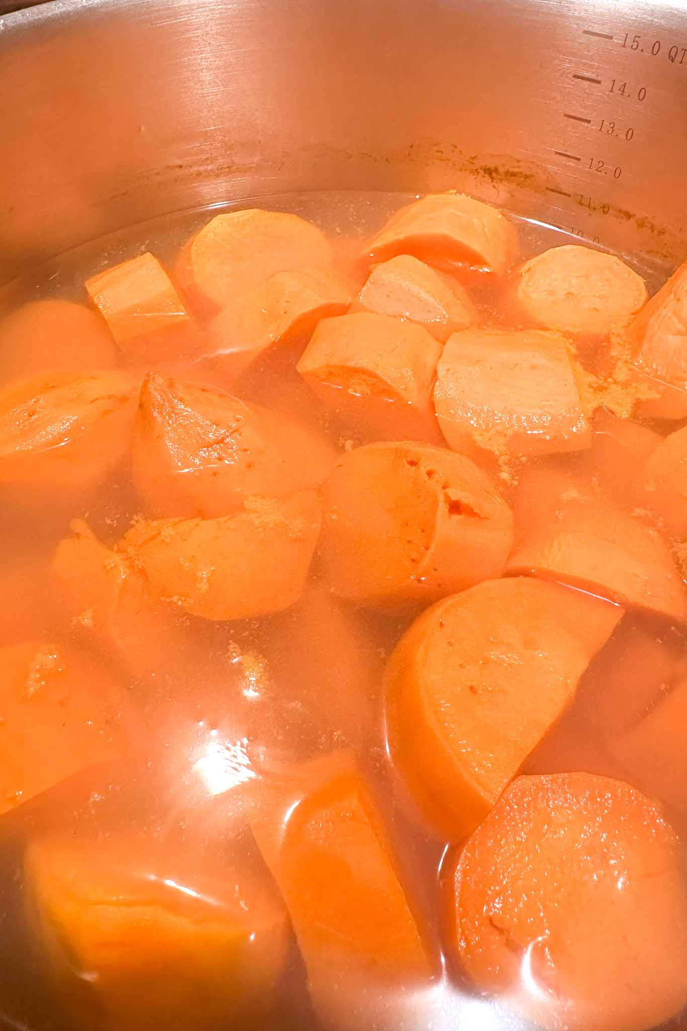 Chunks of orange sweet potatoes boiling in a stainless steel pot filled with water. The pot has measurement markings visible on the inside.