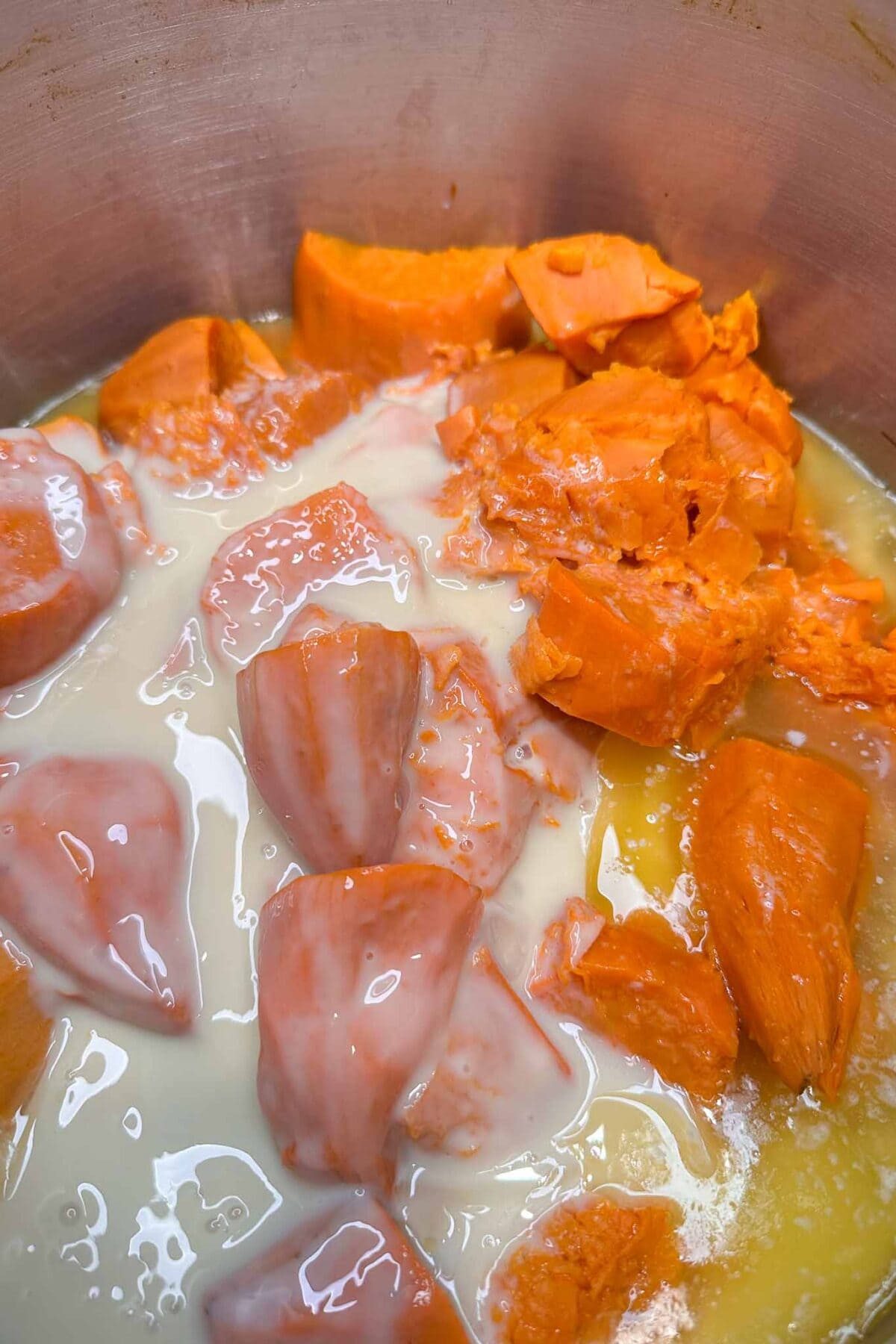 Chunks of orange sweet potato with a creamy liquid, possibly sweetened condensed milk, in a metal pot, ready to be mixed or cooked.