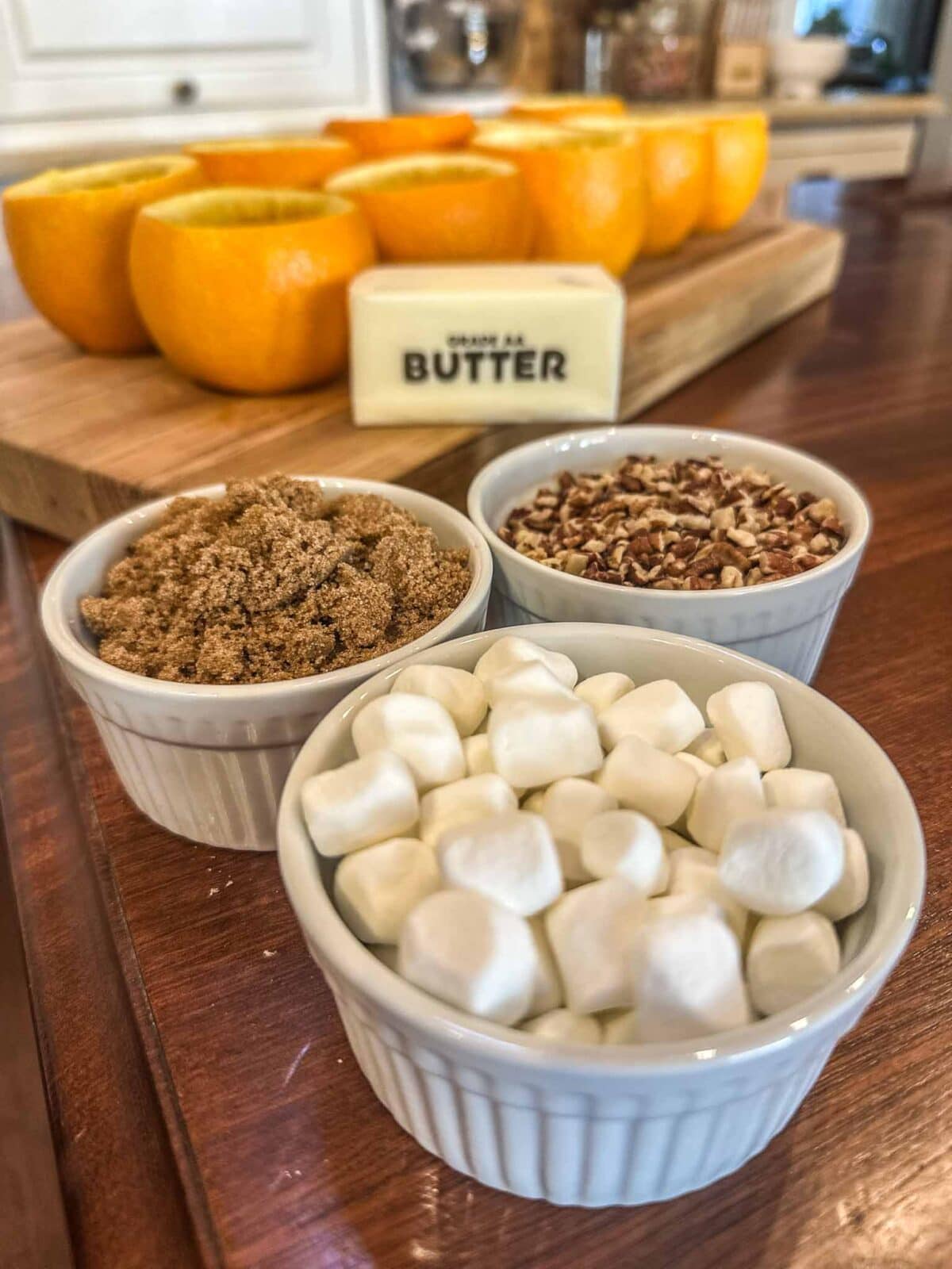 Three small ramekins filled with mini marshmallows, brown sugar, and chopped nuts sit on a wooden counter. Behind them are halved oranges and a stick of butter on a cutting board.