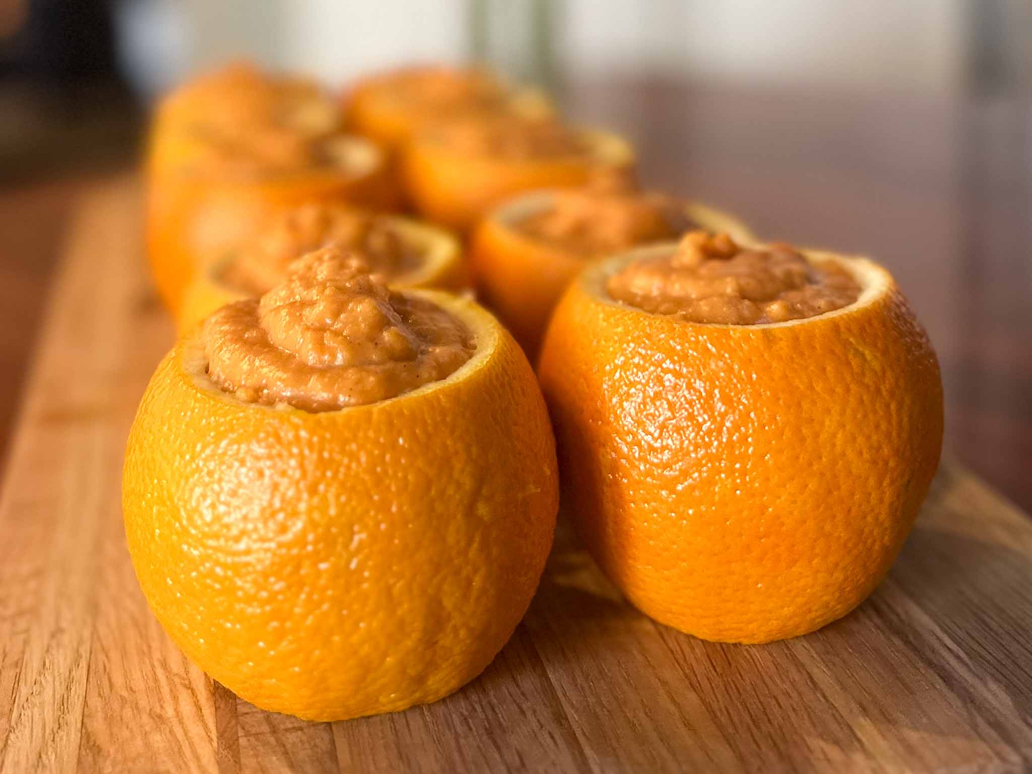 Several hollowed-out oranges filled with a creamy orange mixture are arranged in a row on a wooden surface, with the focus on the oranges in the front.