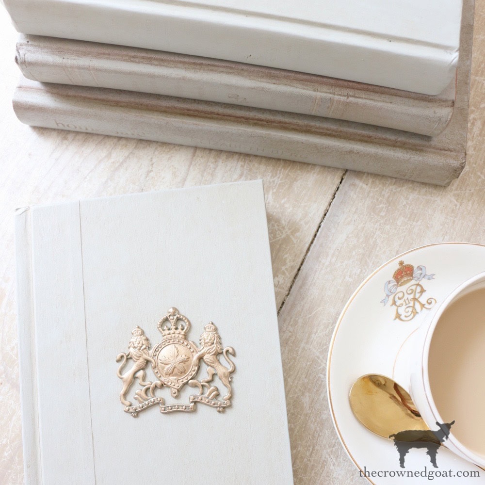 A stack of pale, vintage books beside a white teacup filled with tea and a gold spoon on a matching saucer, set on a light wooden surface. The top book features a royal crest on its cover.