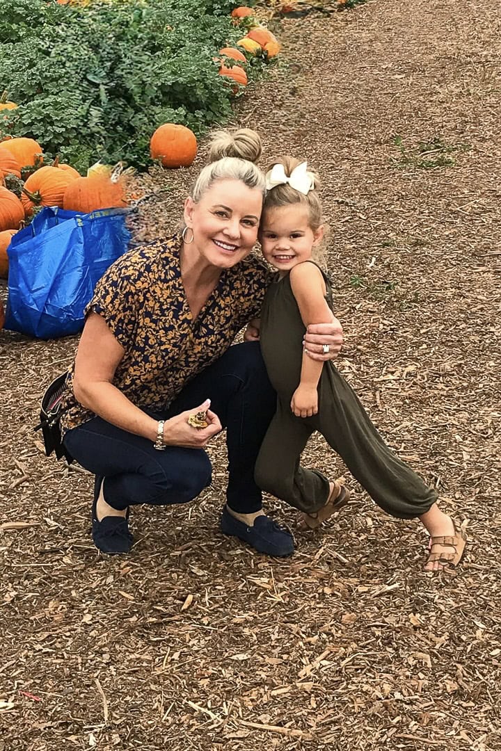 A woman and a young girl smile and pose together at a pumpkin patch with pumpkins, drying pumpkin stems, and a blue bag in the background. The ground is covered with mulch, and the girl wears a green outfit with a bow in her hair.