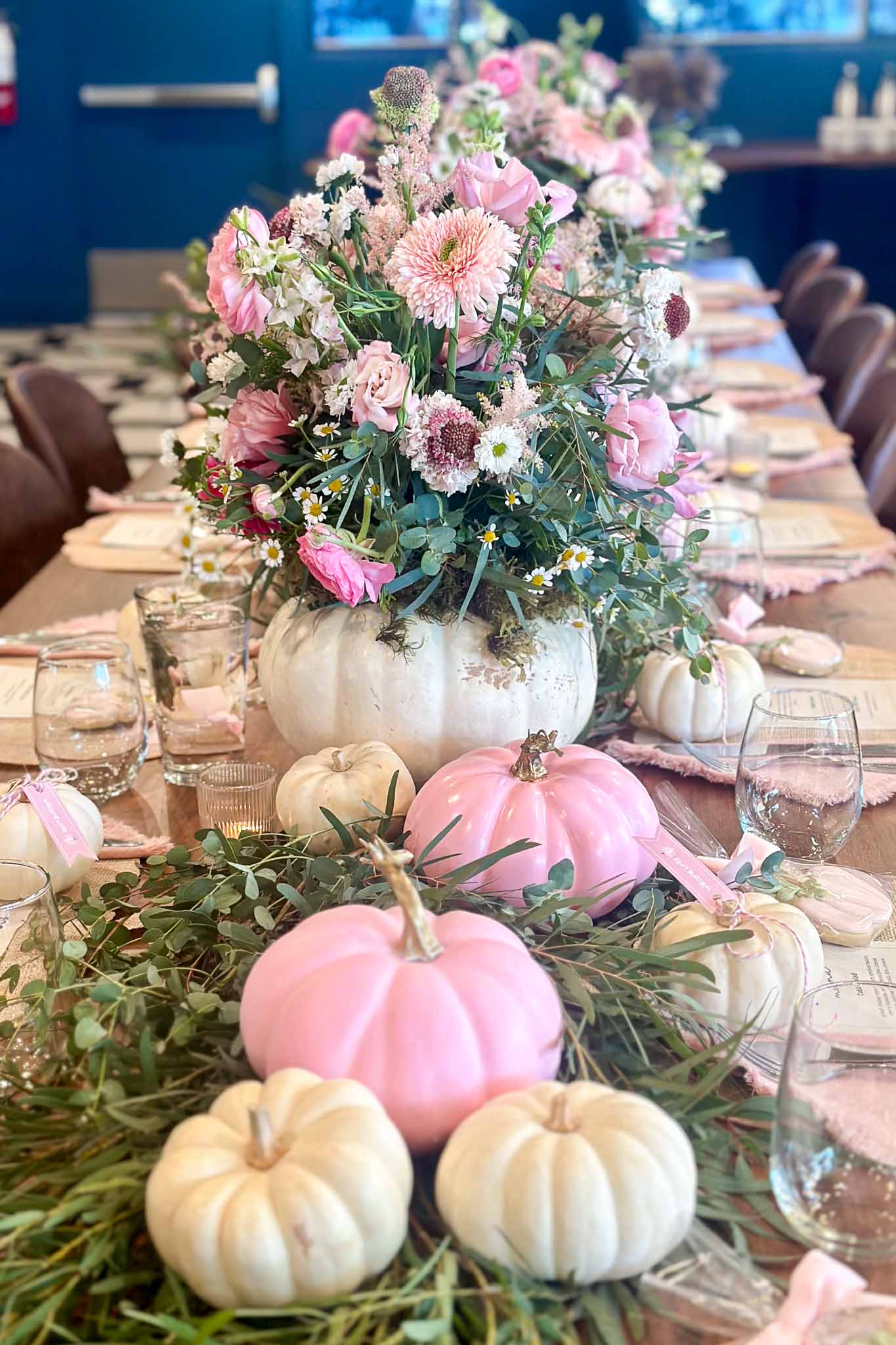 A festive table centerpiece features white and pink pumpkins surrounded by greenery, with large white pumpkins used as vases for pink and white flowers, creating a soft and elegant autumn-themed display.