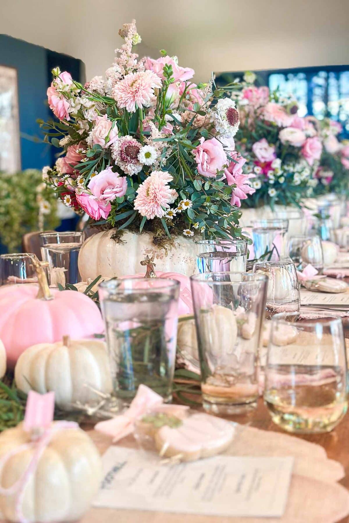 A decorated table features white and pink pumpkins, glassware, menus, and centerpieces of pink and white flowers arranged in white pumpkins, creating a soft, elegant autumn theme.