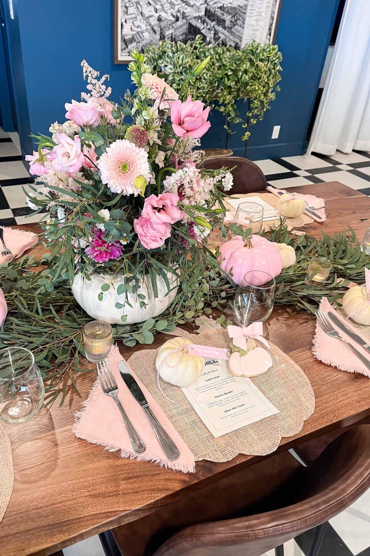A wooden dining table set with pink flowers in a white vase, greenery, small white pumpkins, glassware, cutlery, pink napkins, and printed menus. The background features a blue wall and black-and-white checkered floor.