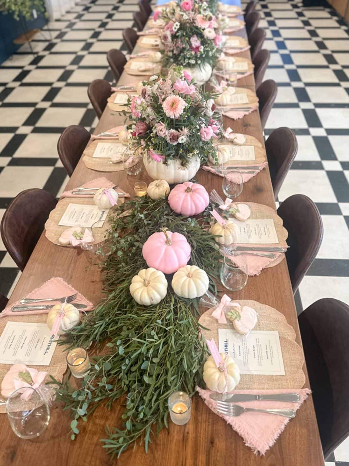 A long wooden table set for a meal features pink and white pumpkins, greenery, flowers in pumpkin vases, menus, pink napkins, and small candles, with a black and white checkered floor beneath.