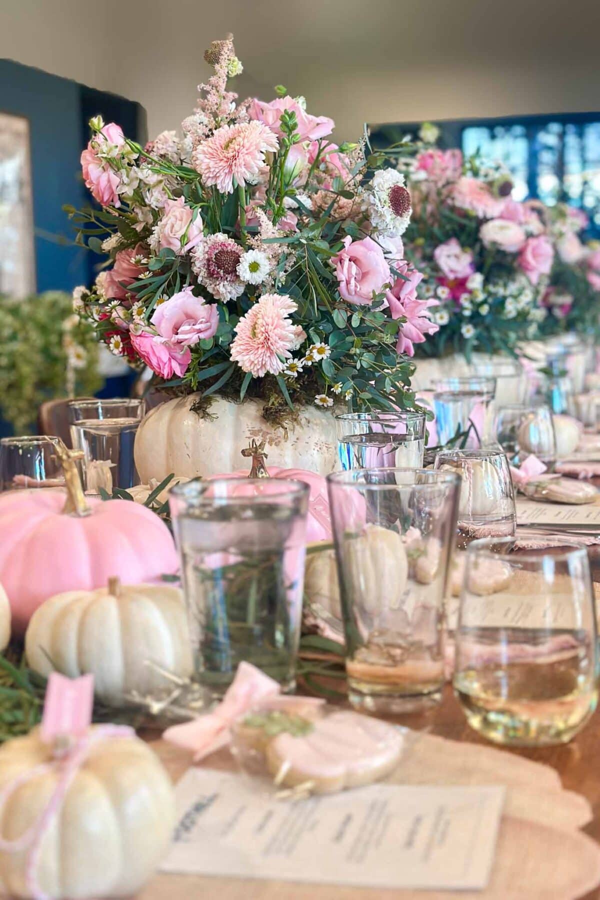 A festive table setting features white and pink pumpkins, glasses, menus, and a centerpiece bouquet of pink flowers arranged in a white pumpkin. The overall decor has a soft, elegant autumn theme.