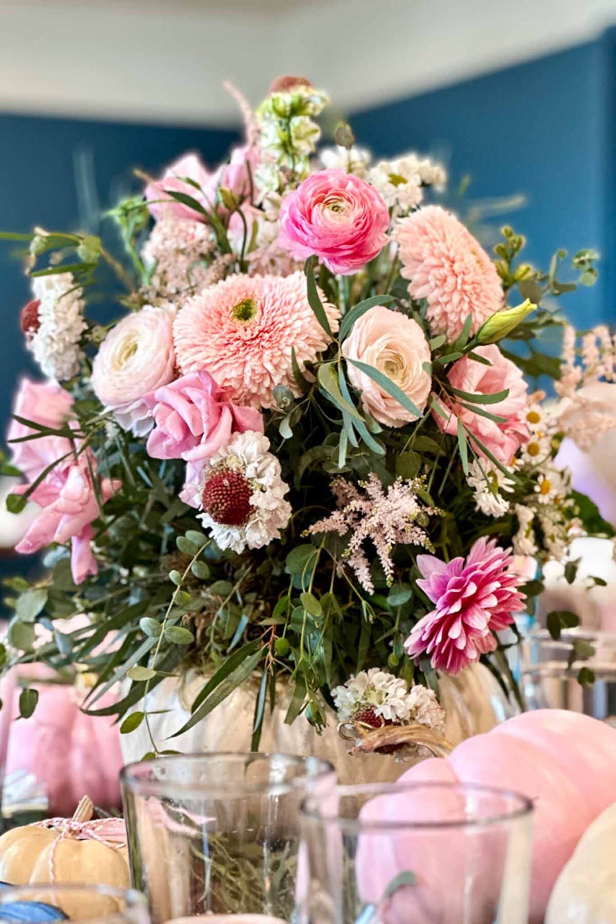 A bouquet of pink and white flowers, including roses and dahlias, arranged in a white pumpkin vase. The arrangement is surrounded by glassware and pastel pink pumpkins on a table with a blue background.