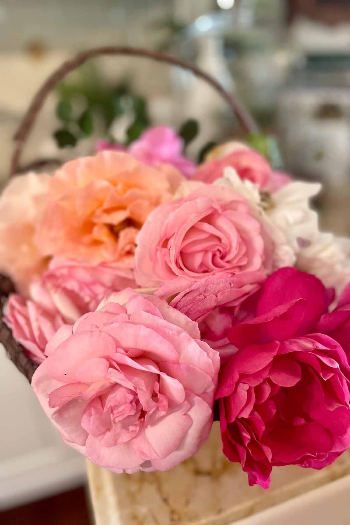 A basket filled with various shades of pink and peach roses, with delicate petals and a few white flowers, sits on a marble surface in a softly lit indoor setting.