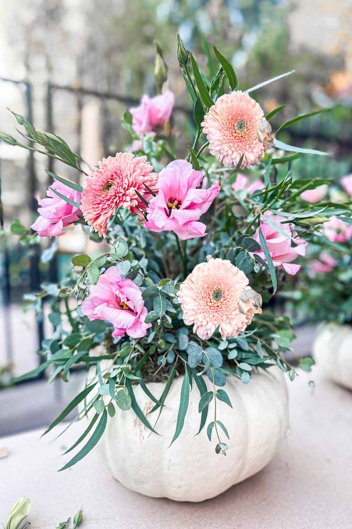 A white pumpkin is used as a vase, holding a floral arrangement of pink gerbera daisies, lisianthus, and green foliage on a light outdoor surface.