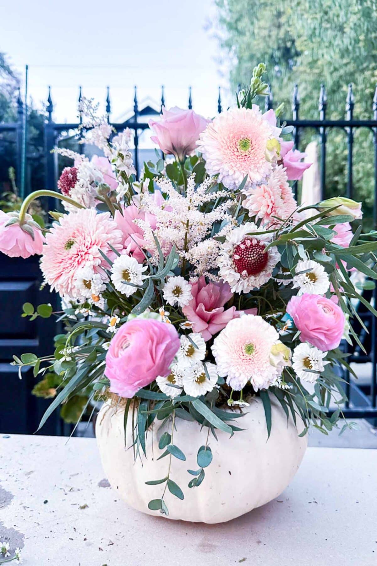 A white pumpkin is used as a vase, holding an arrangement of pink, white, and light purple flowers with greenery, set outdoors in front of a black iron fence and green trees.