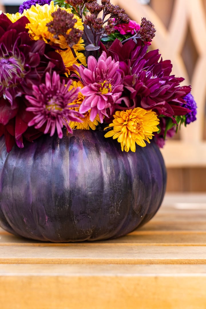 A dark purple pumpkin is transformed into a Dried Flower Pumpkin Centerpiece, holding a vibrant bouquet of yellow, purple, and magenta flowers atop a wooden surface with a softly blurred background.