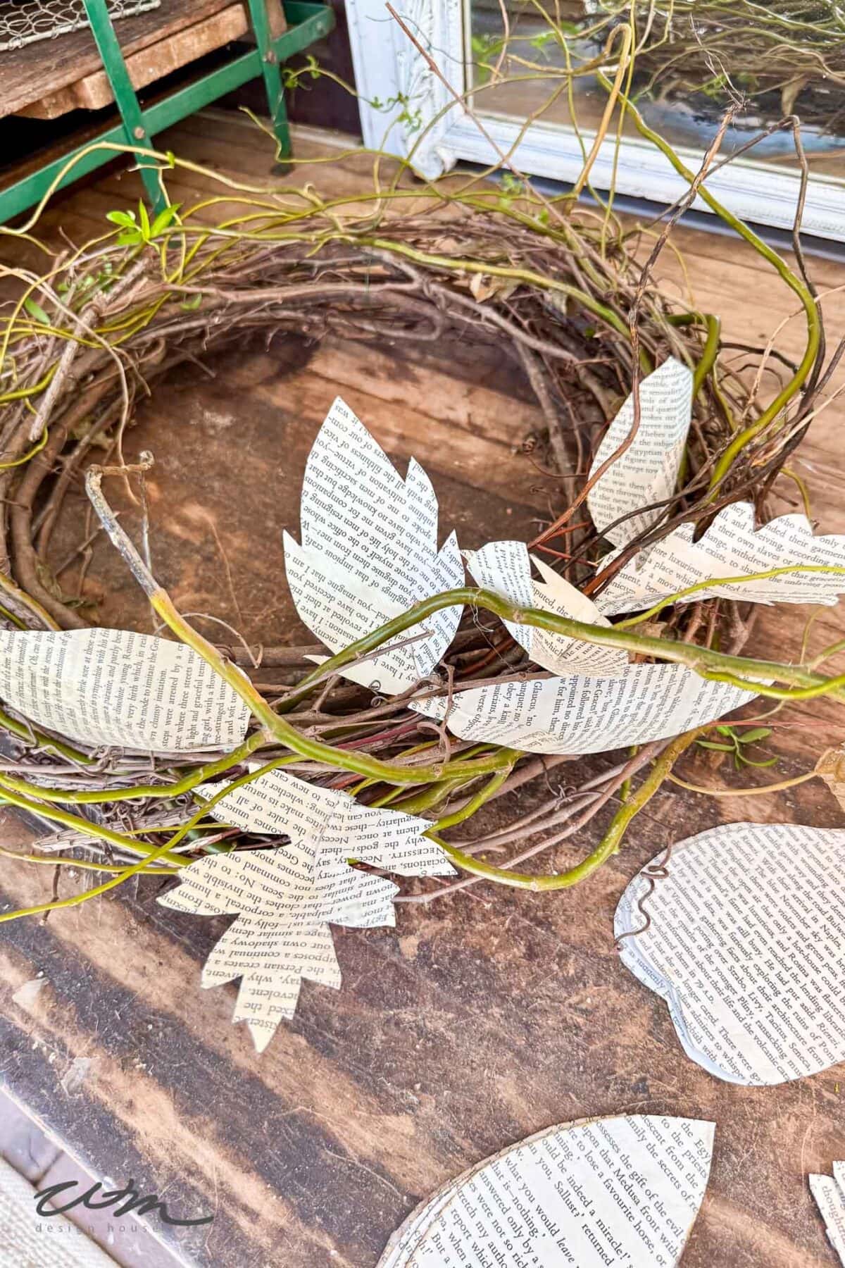A rustic grapevine wreath decorated with leaves cut from book pages rests on a wooden surface. Additional paper leaves are scattered nearby.