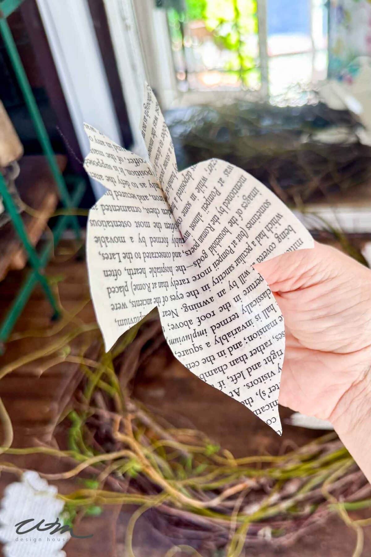A hand holding a butterfly-shaped cutout made from printed book pages, with a wooden surface and green plants in the background.