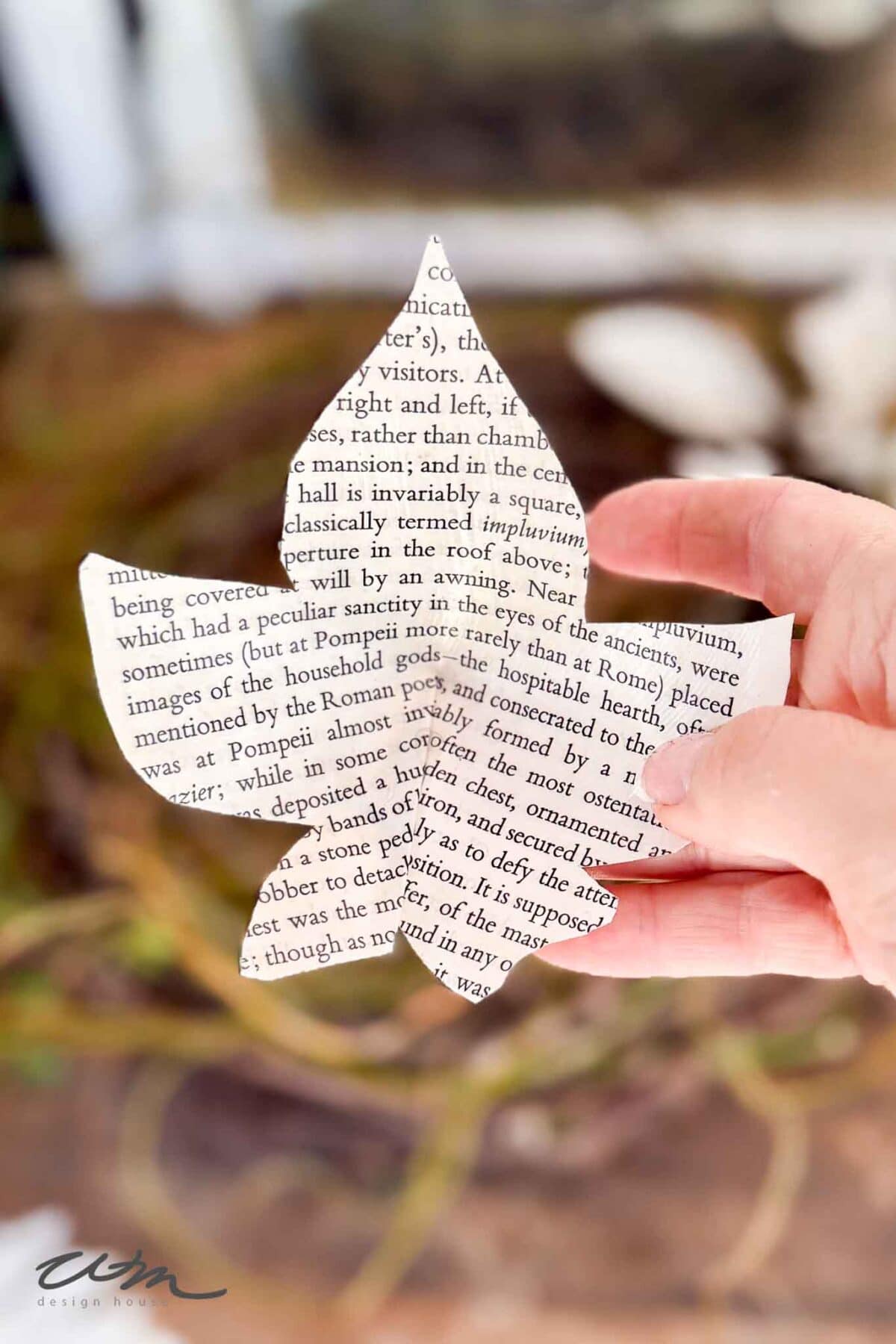 A hand holds a paper cutout shaped like a leaf, made from a printed page with black text. The background is blurred with earthy tones, suggesting plants or foliage.