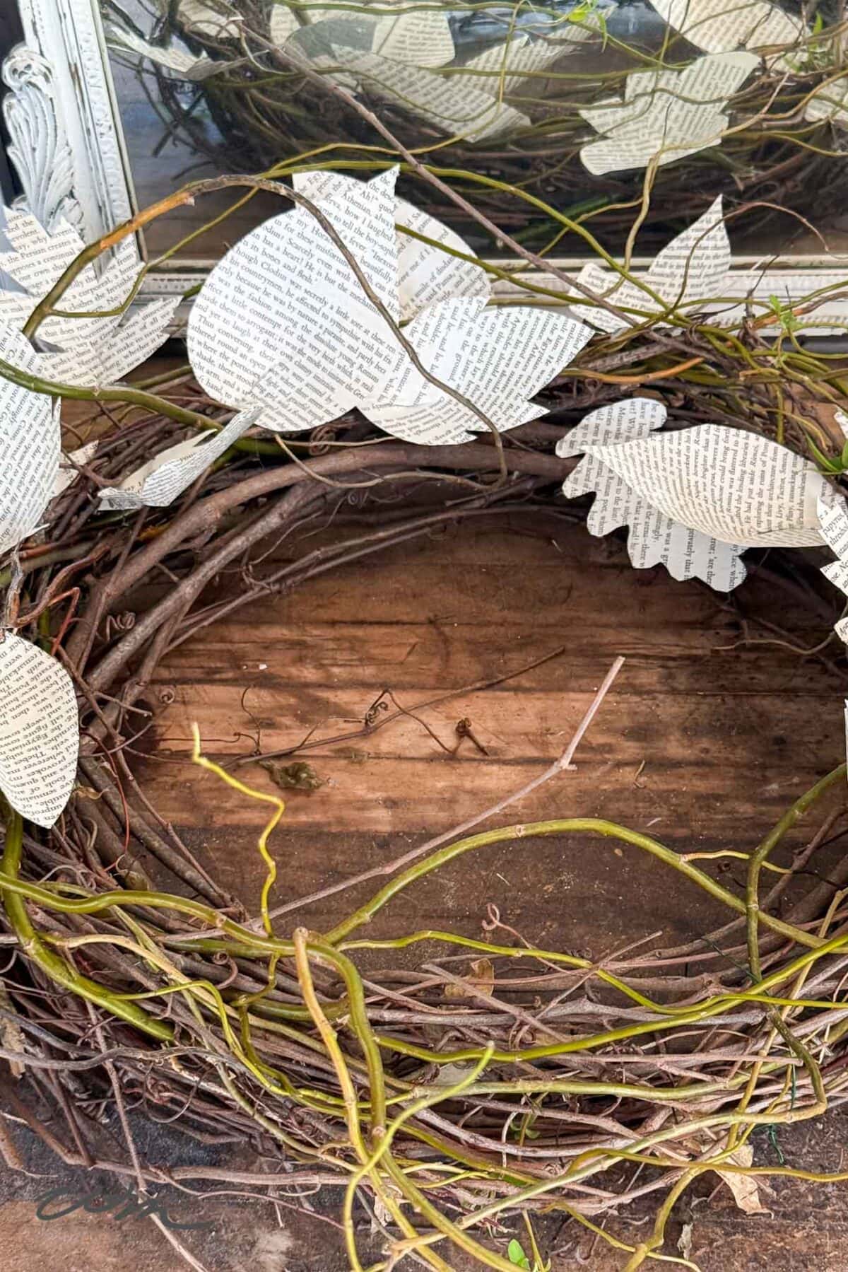 A wreath made of intertwined brown vines with paper leaves cut from book pages, resting on a wooden surface near a mirror.