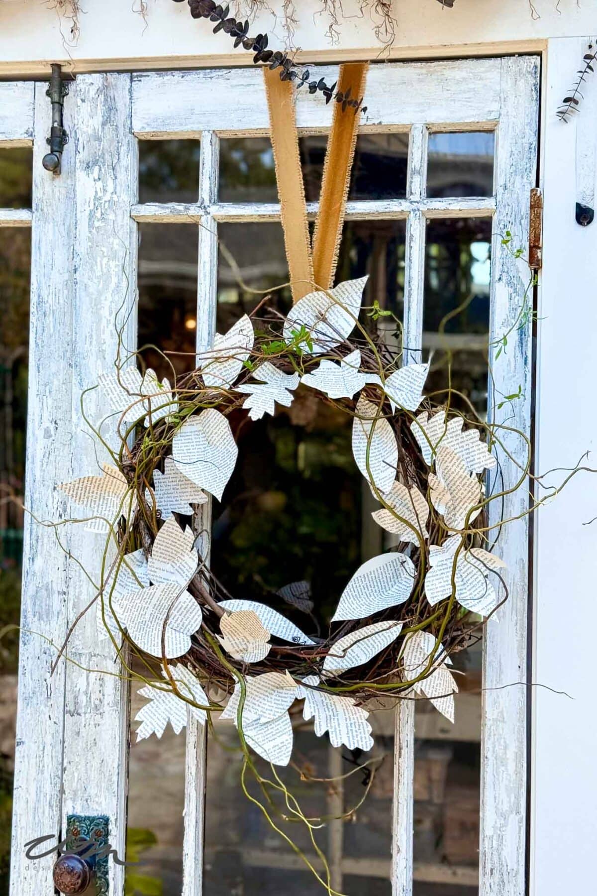 A rustic grapevine wreath decorated with paper leaves hangs from a tan ribbon on a weathered, white-painted door with glass panels.