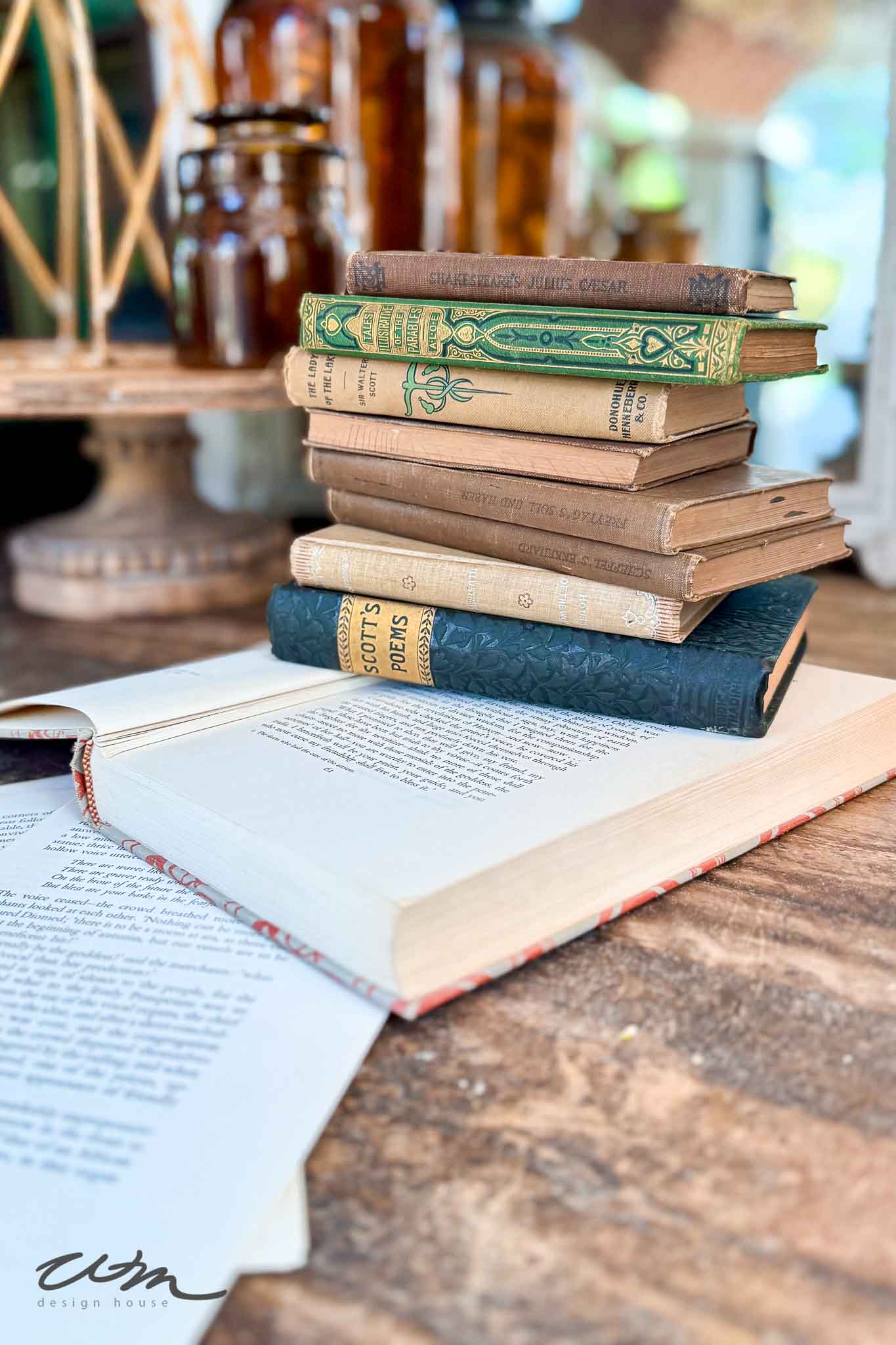 A stack of vintage books sits on a rustic wooden table, with an open book in front. Glass jars and shelves are blurred in the background, creating a cozy, antique atmosphere.