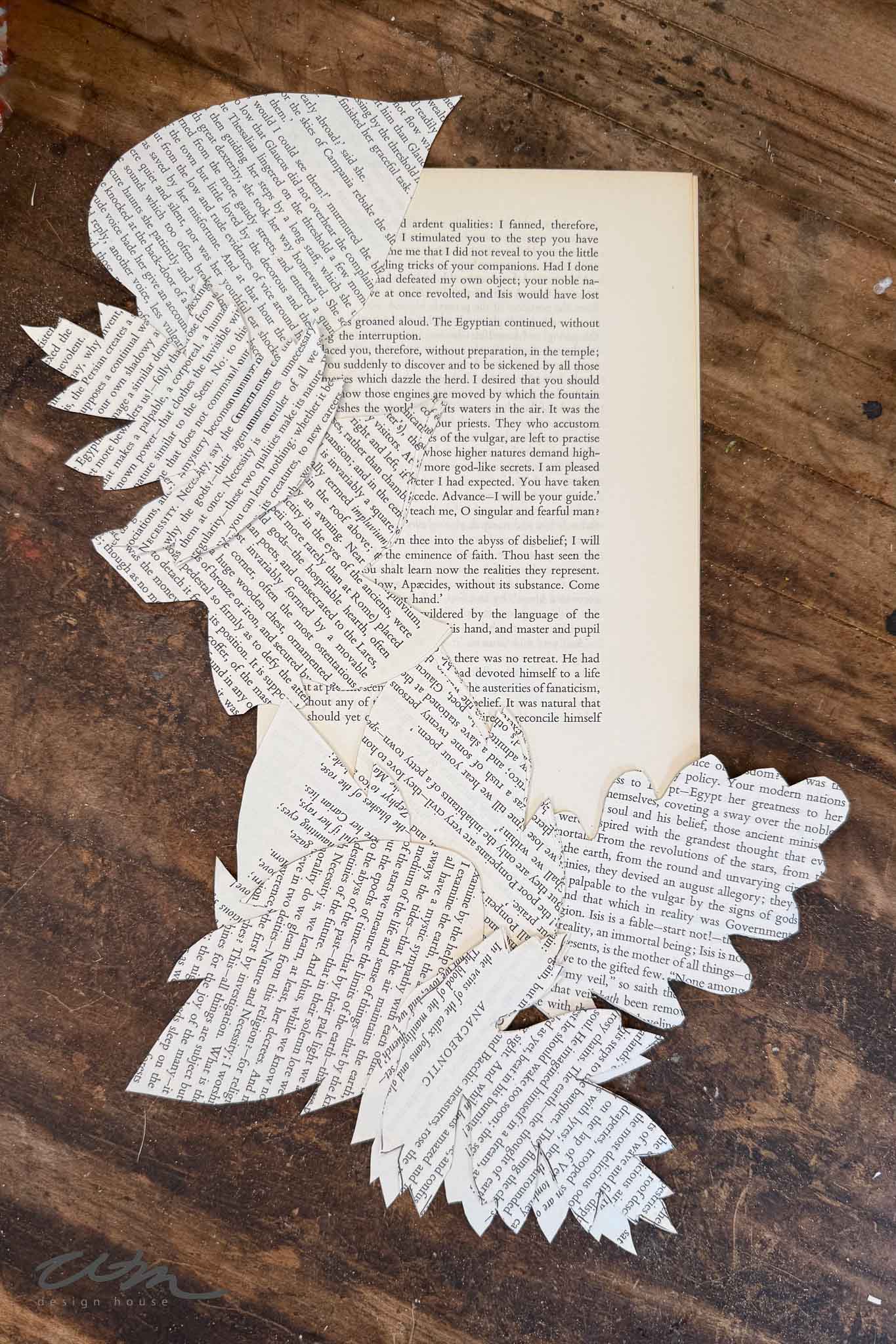 A book page lies open on a wooden surface, with intricate leaf and feather shapes cut from book pages arranged artistically on top, partially covering the book.