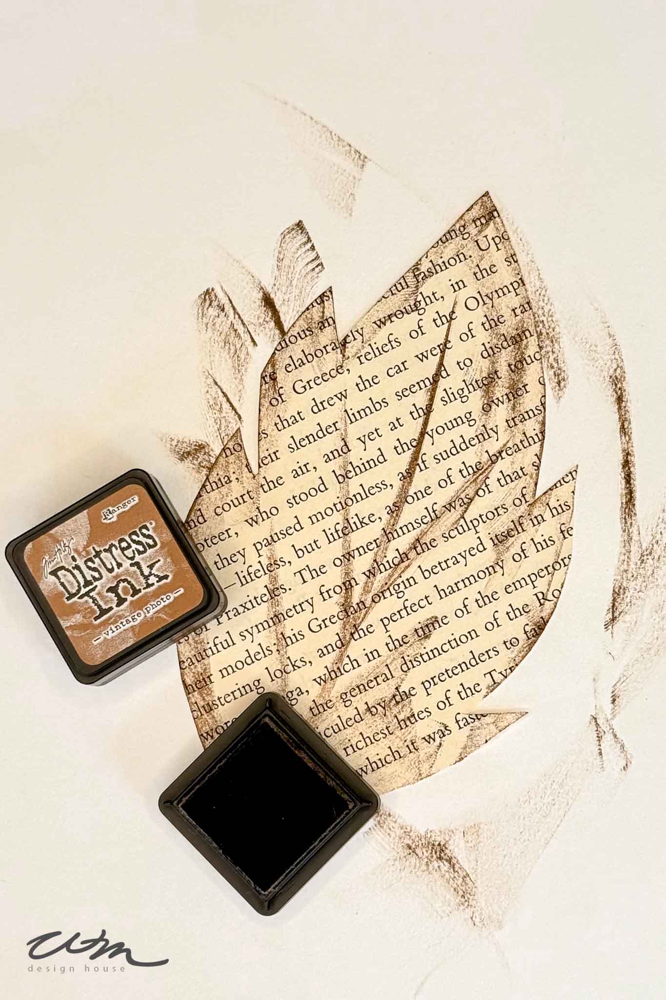 A leaf shape cut out of text-covered paper is outlined and shaded with brown ink. Two Distress Ink pads, one open and one closed, are placed near the leaf on a cream-colored background.