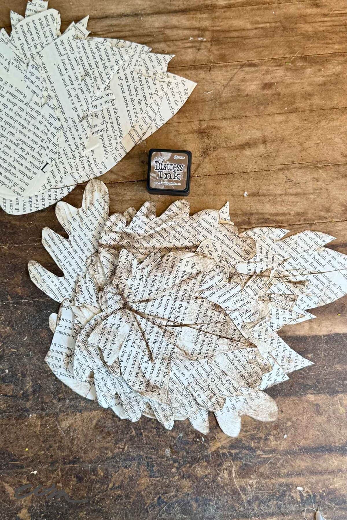 Paper leaves cut from book pages are arranged on a wooden surface, with a brown Distress Ink pad placed above them. The leaves are layered and have text printed on them.