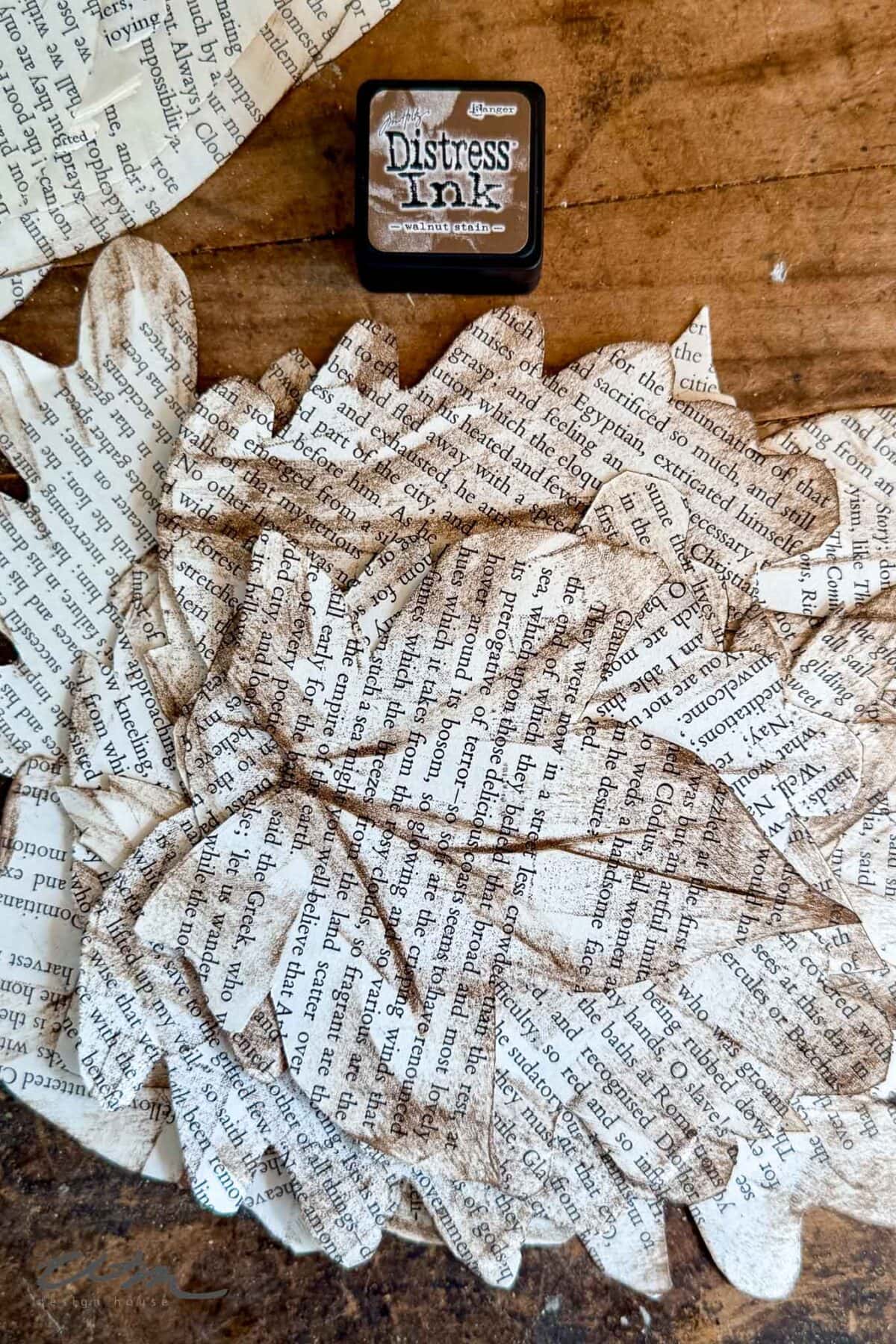 Several paper leaves cut from book pages are scattered on a wooden surface, with a small ink pad labeled "Distress Ink" positioned nearby. The leaves have darkened, inked edges for a vintage effect.