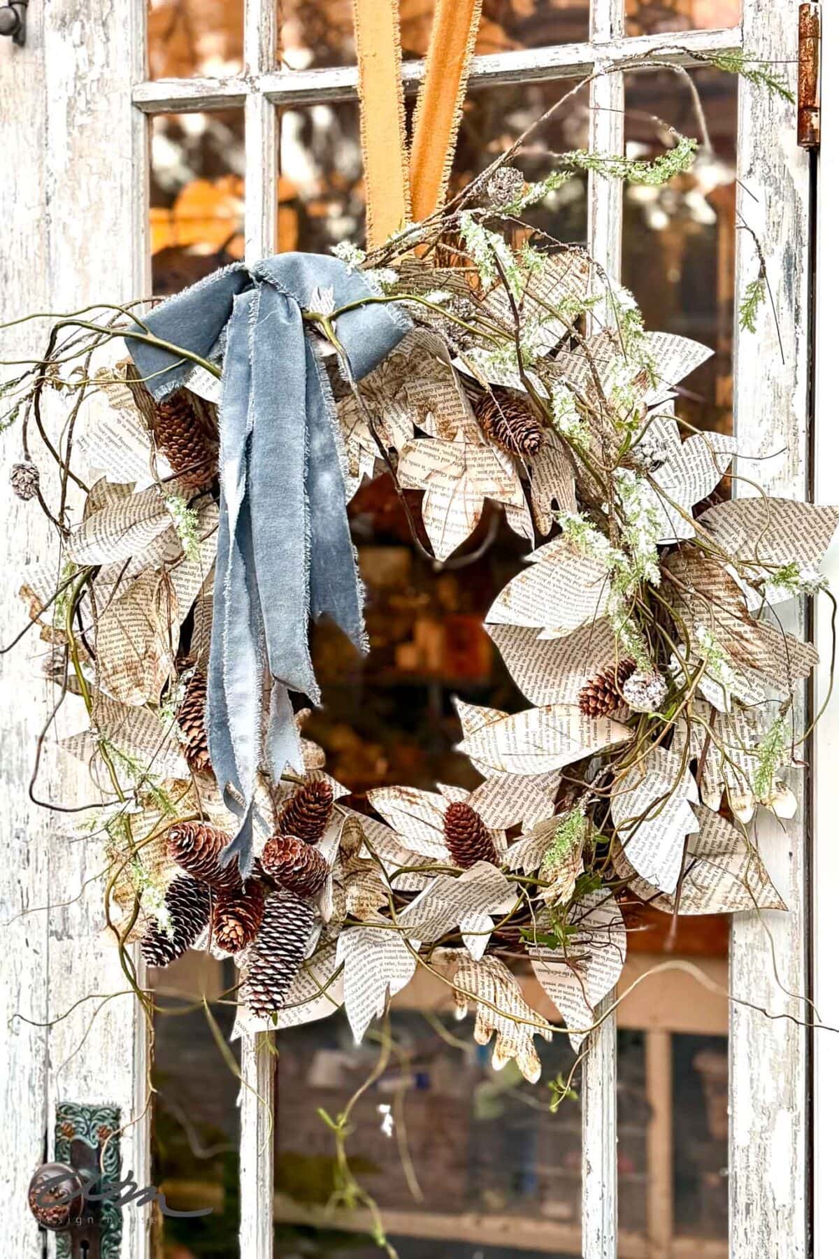 A rustic wreath made of pinecones, dried branches, and paper leaves with text, adorned with a blue velvet ribbon, hangs on a weathered glass door.