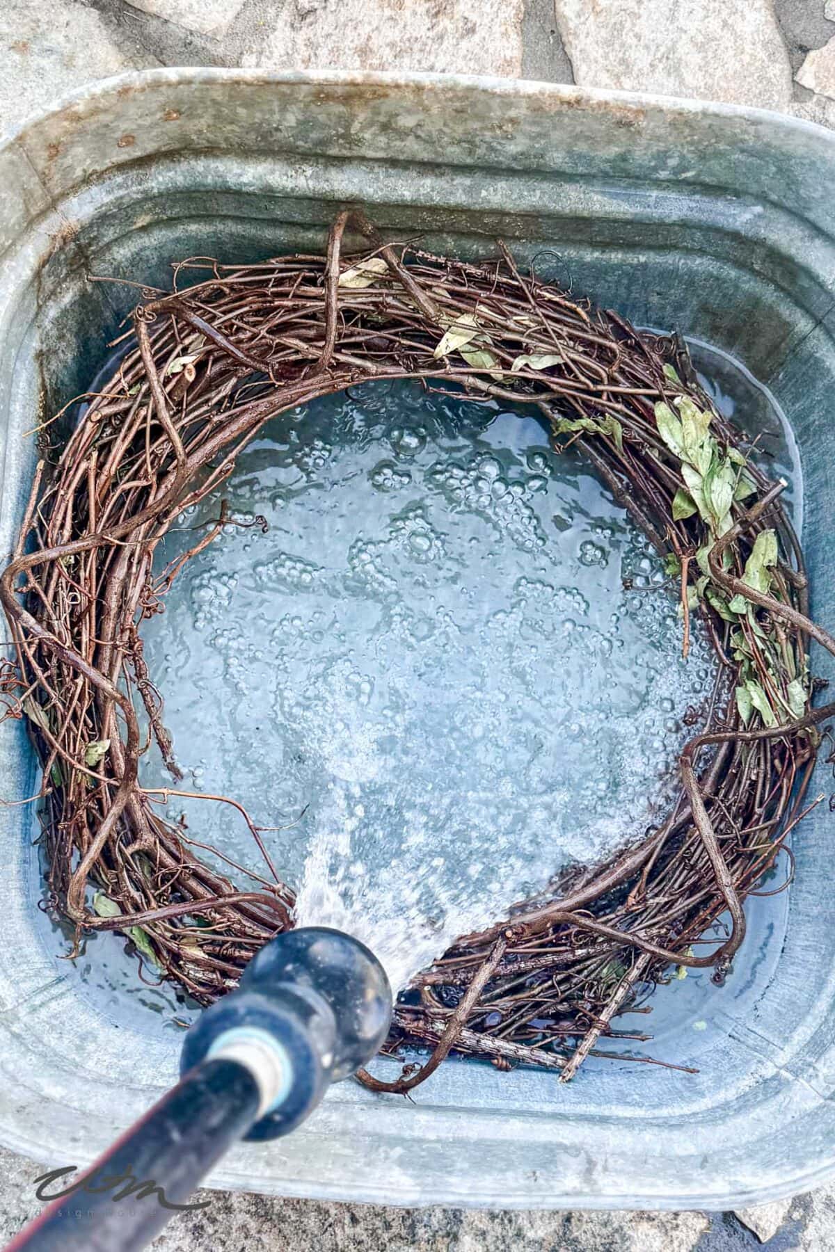 A grapevine wreath is being washed with water from a hose inside a metal basin. The wreath floats in clear water, and a few green leaves remain attached to the twigs. Stone pavers are visible in the background.