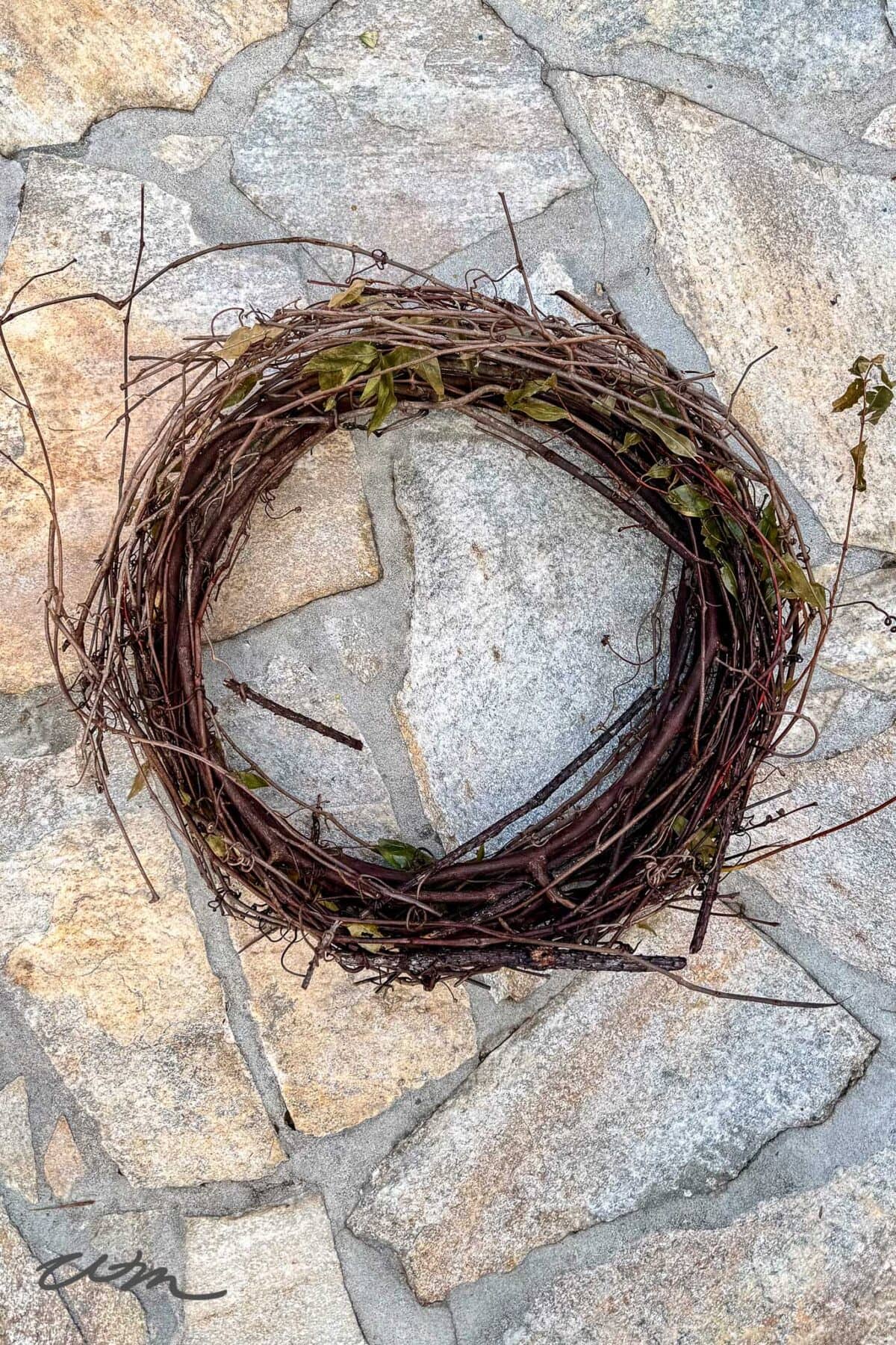 A circular wreath made of intertwined brown vines and twigs, with some small green leaves and subtle accents reminiscent of a book page wreath, lies on a light gray stone pavement.