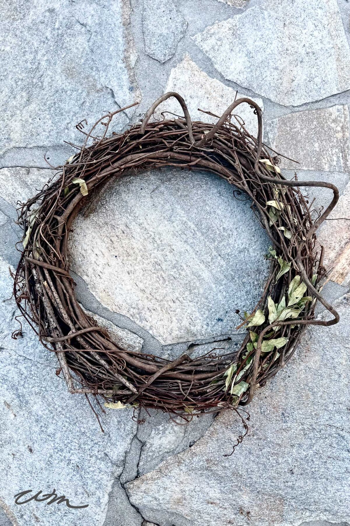 A simple grapevine wreath, reminiscent of a book page wreath, is decorated with a few dried leaves and hangs on a light gray stone wall.