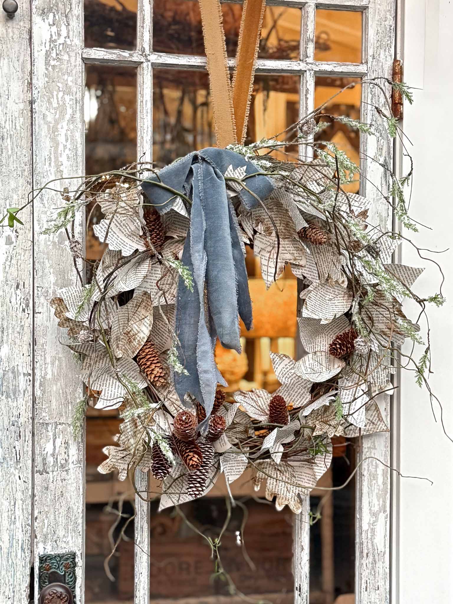 A rustic book page wreath made of pinecones, twigs, and paper leaves with printed text hangs on a weathered glass door, adorned with a blue fabric ribbon and a tan knotted strap.
