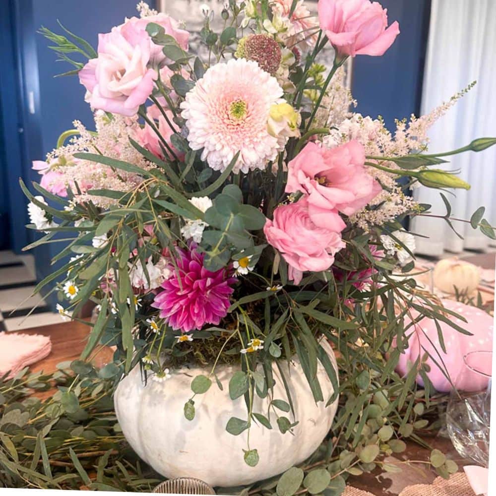 A white ceramic vase filled with a lush White Pumpkin Flower Arrangement of pink roses, daisies, and greenery sits on a decorated table, creating a fresh and elegant centerpiece.