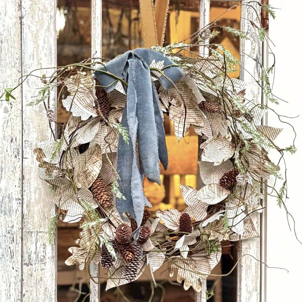 A rustic book page wreath made from paper leaves, pinecones, and twigs, adorned with a blue velvet ribbon, hangs on a weathered wooden door with a glass panel. Warm interior lights glow softly in the background.