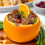 Stuffed orange bowl filled with savory stuffing, topped with herbs and an orange slice, served on a white plate with green beans and sauce. Cranberry sauce and meat are in the background.