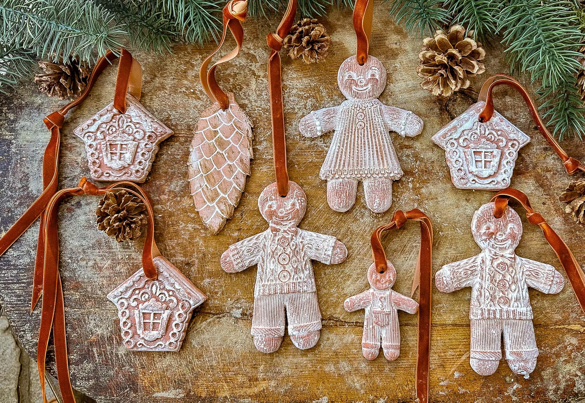 Assorted gingerbread-themed ornaments, including houses, pinecones, and smiling gingerbread people, are decorated with white icing details—perfect inspiration if you’re looking for how to make air dry clay ornaments at home.