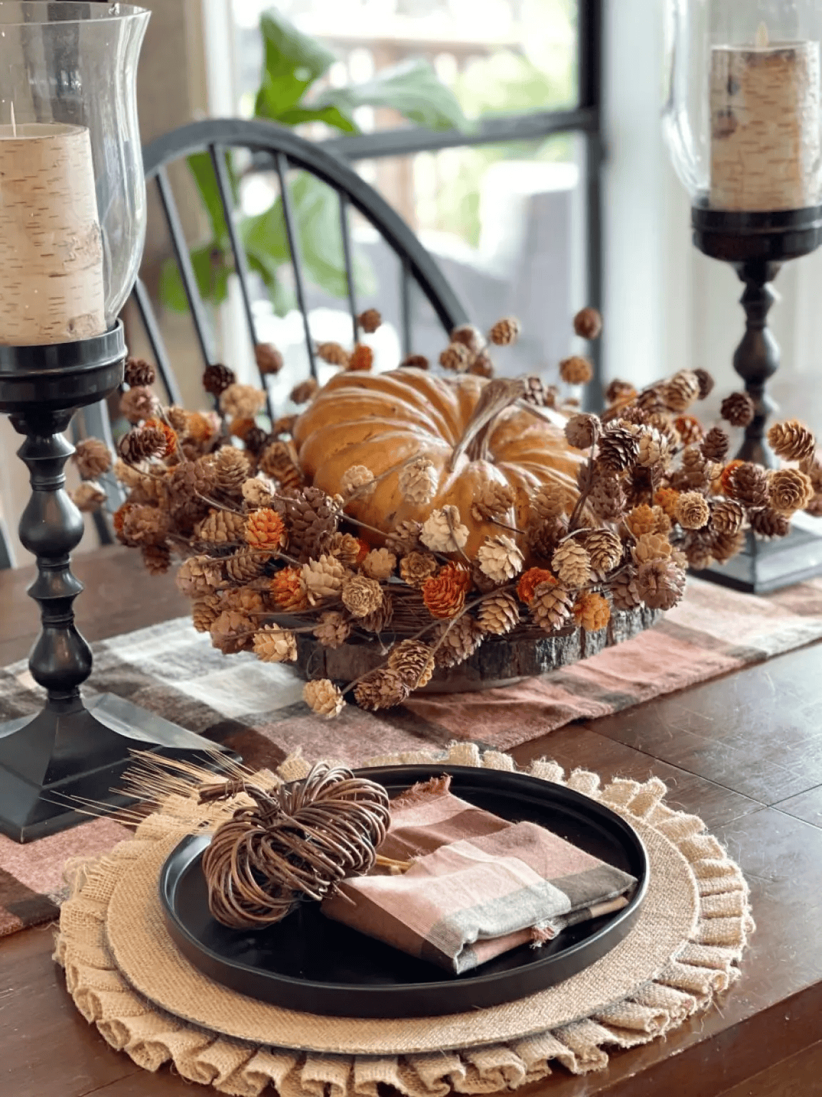 A fall-themed dining table features a Dried Flower Pumpkin Centerpiece with pinecones on a wooden table. Nearby, a black plate with a rustic napkin and twig pumpkin rests on a fringed placemat between two candle holders.