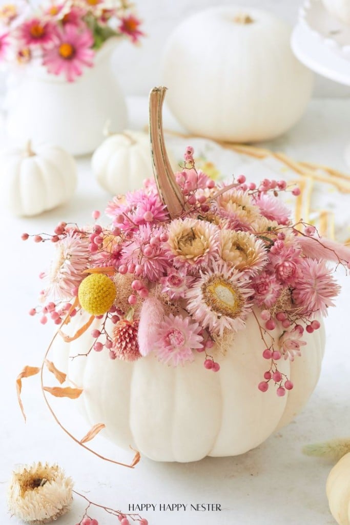 A white pumpkin serves as a vase for this stunning Dried Flower Pumpkin Centerpiece, filled with pink, yellow, and cream dried flowers and berries. More white pumpkins and flowers are softly blurred in the background on a white surface.