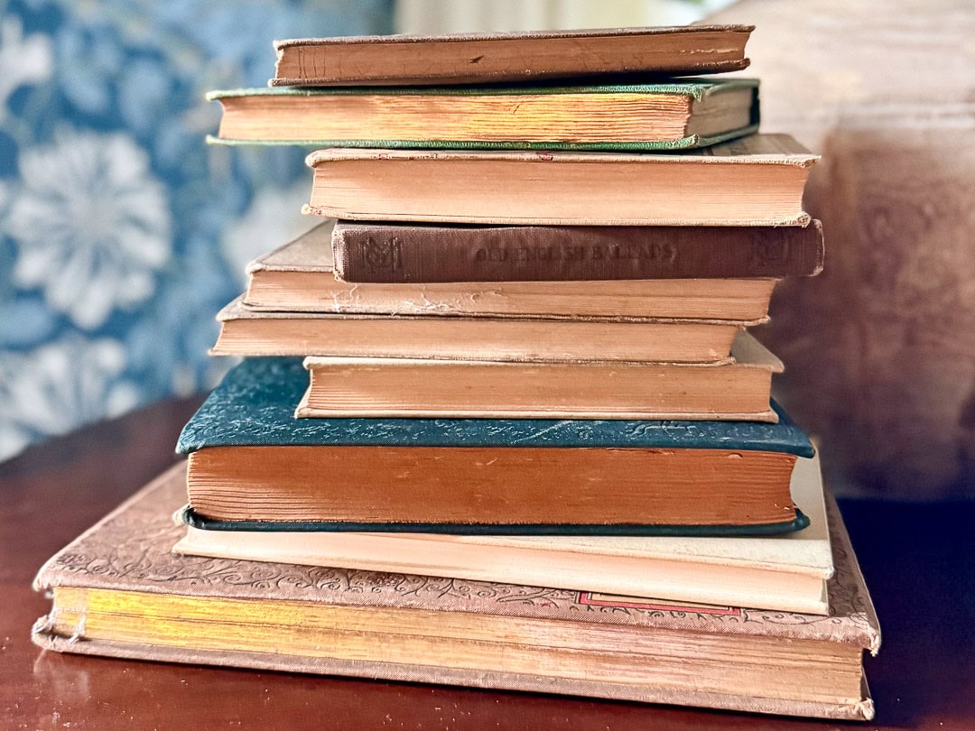 A stack of old, worn books with various colored covers sits on a wooden surface, exuding vintage charm against a blue floral-patterned background that peeks through.