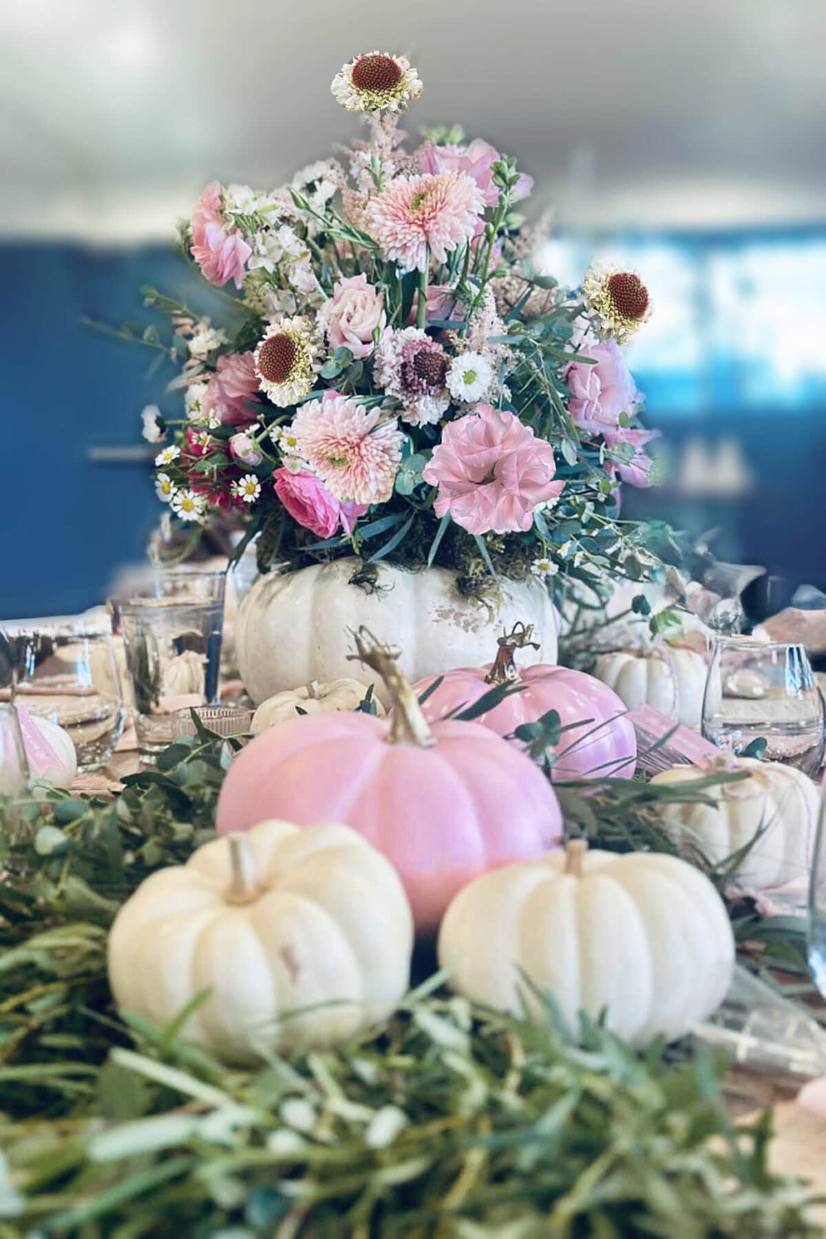 A White Pumpkin Flower Arrangement serves as the centerpiece, filled with pink and white blooms, surrounded by small pastel pumpkins, green foliage, and elegant glassware on a beautifully decorated table.