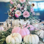 A White Pumpkin Flower Arrangement serves as the centerpiece, filled with pink and white blooms, surrounded by small pastel pumpkins, green foliage, and elegant glassware on a beautifully decorated table.
