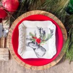 A festive table setting features a red plate and square white plate with a reindeer whose antlers are adorned with leaves and red flowers. The plates rest on DIY sheet music charger plates, surrounded by pine branches and red ornaments.