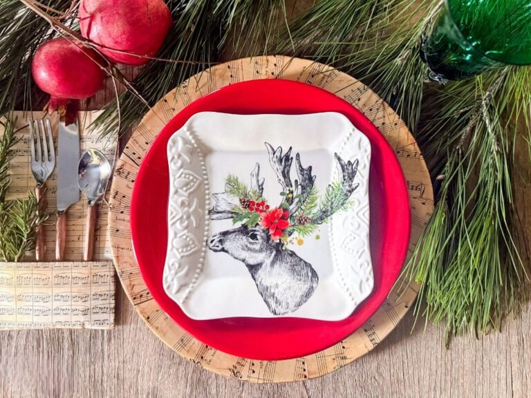 A festive table setting features a red plate and square white plate with a reindeer whose antlers are adorned with leaves and red flowers. The plates rest on DIY sheet music charger plates, surrounded by pine branches and red ornaments.