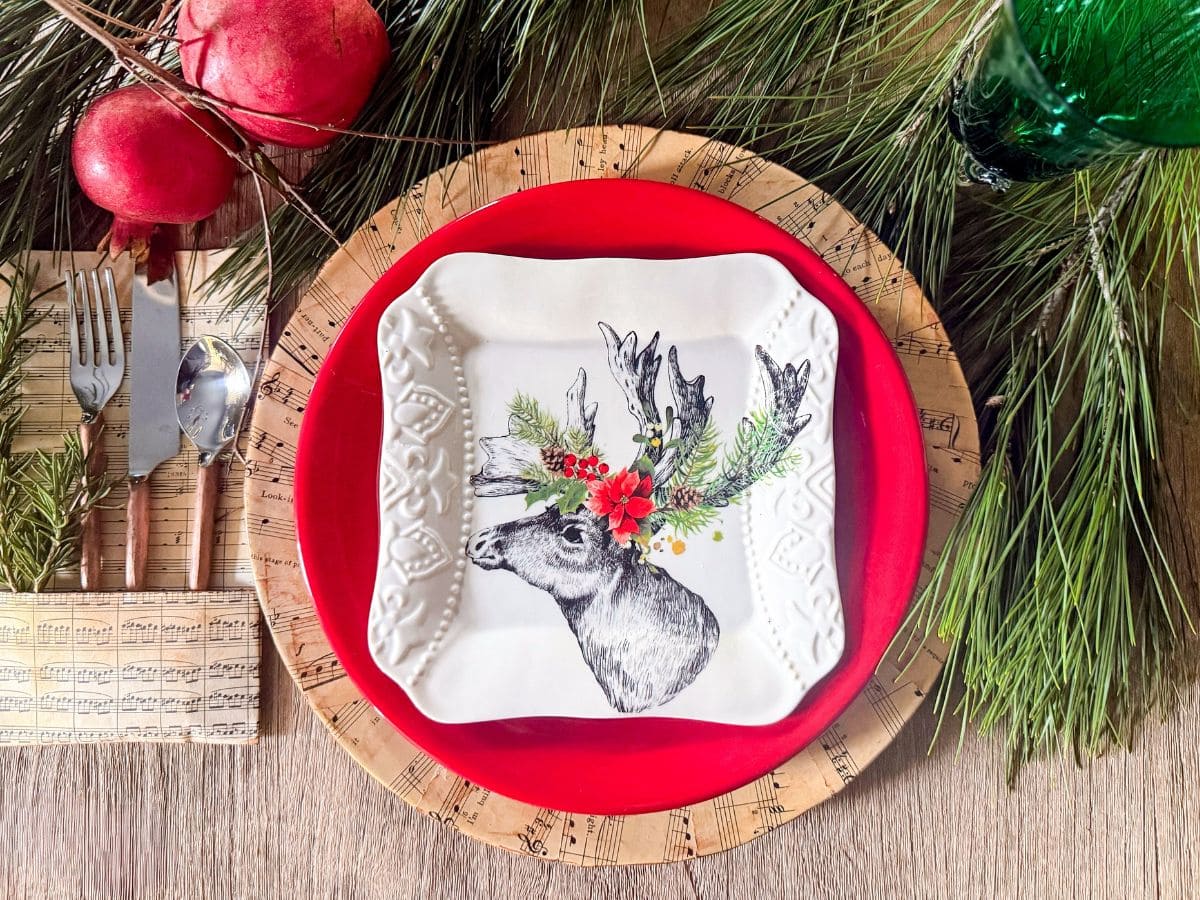 A festive table setting features a red plate and square white plate with a reindeer whose antlers are adorned with leaves and red flowers. The plates rest on DIY sheet music charger plates, surrounded by pine branches and red ornaments.