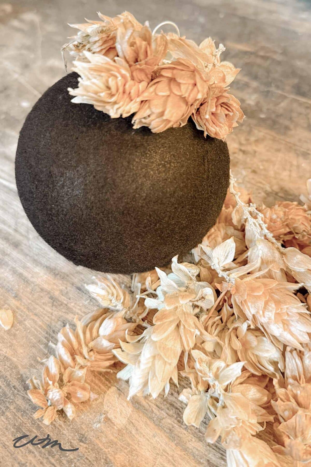A black round ornament decorated with pale dried flowers is displayed on a rustic wooden surface, with additional dried flowers scattered nearby.
